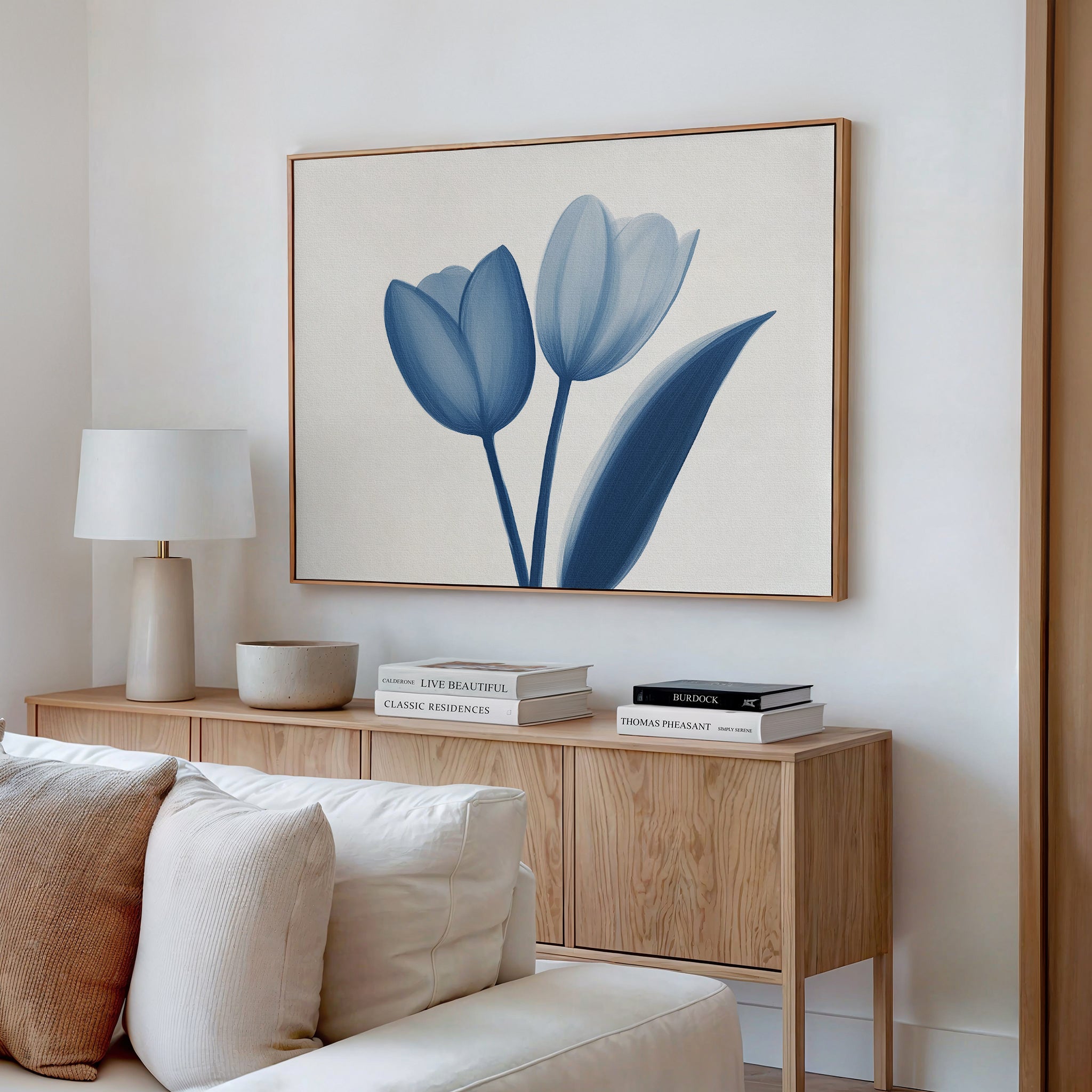 A minimalist living room features a light wood sideboard, neutral decor, and the Cerulean Tulips Canvas Art - Horizontal above. A lamp, bowl, and stacked books lend subtle charm to this Nordic Park-inspired space.