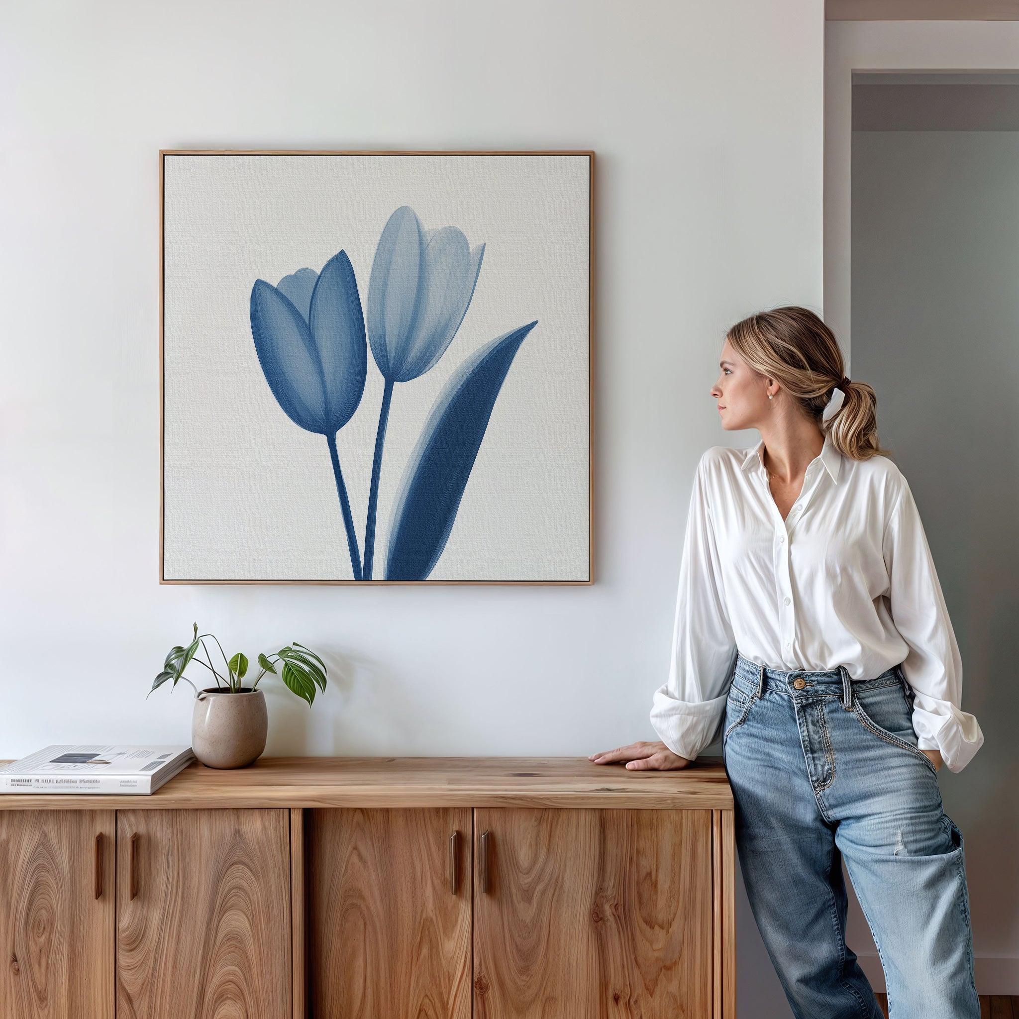 A woman in a white blouse and jeans leans against a wooden cabinet, gazing at the Cerulean Tulips Canvas Art - Square above. A small plant and books decorate the cabinet in this bright, modern, Nordic Park-inspired room.