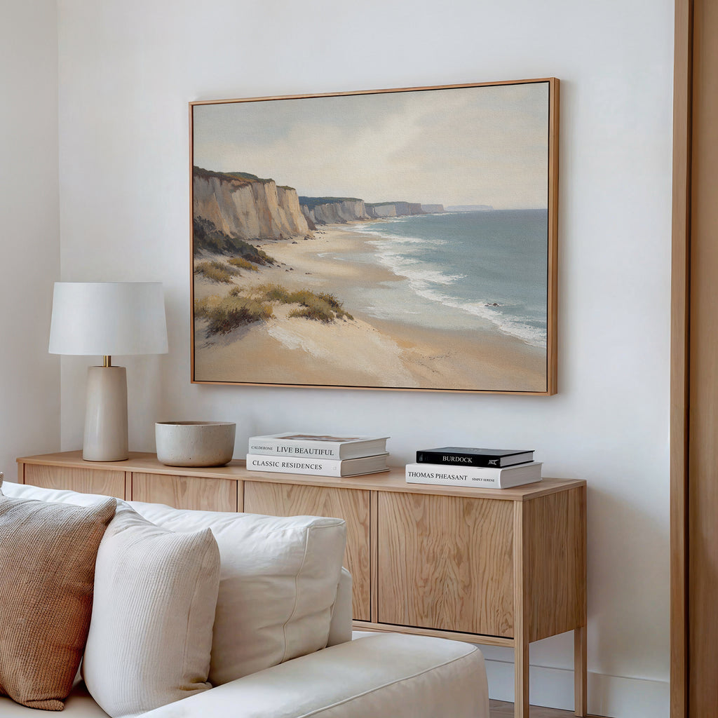 A modern living room features a light wood console with stacked books, a ceramic bowl, lamp, and the Chalk Shores Canvas Art - Horizontal above a cream sofa against a white wall.