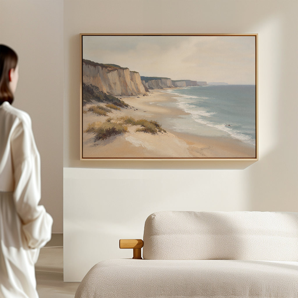 A woman admires the Chalk Shores Canvas Art - Horizontal, featuring sandy cliffs and gentle waves, hanging above a light-colored sofa in a softly lit room.
