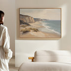 A woman admires the Chalk Shores Canvas Art - Horizontal, featuring sandy cliffs and gentle waves, hanging above a light-colored sofa in a softly lit room.