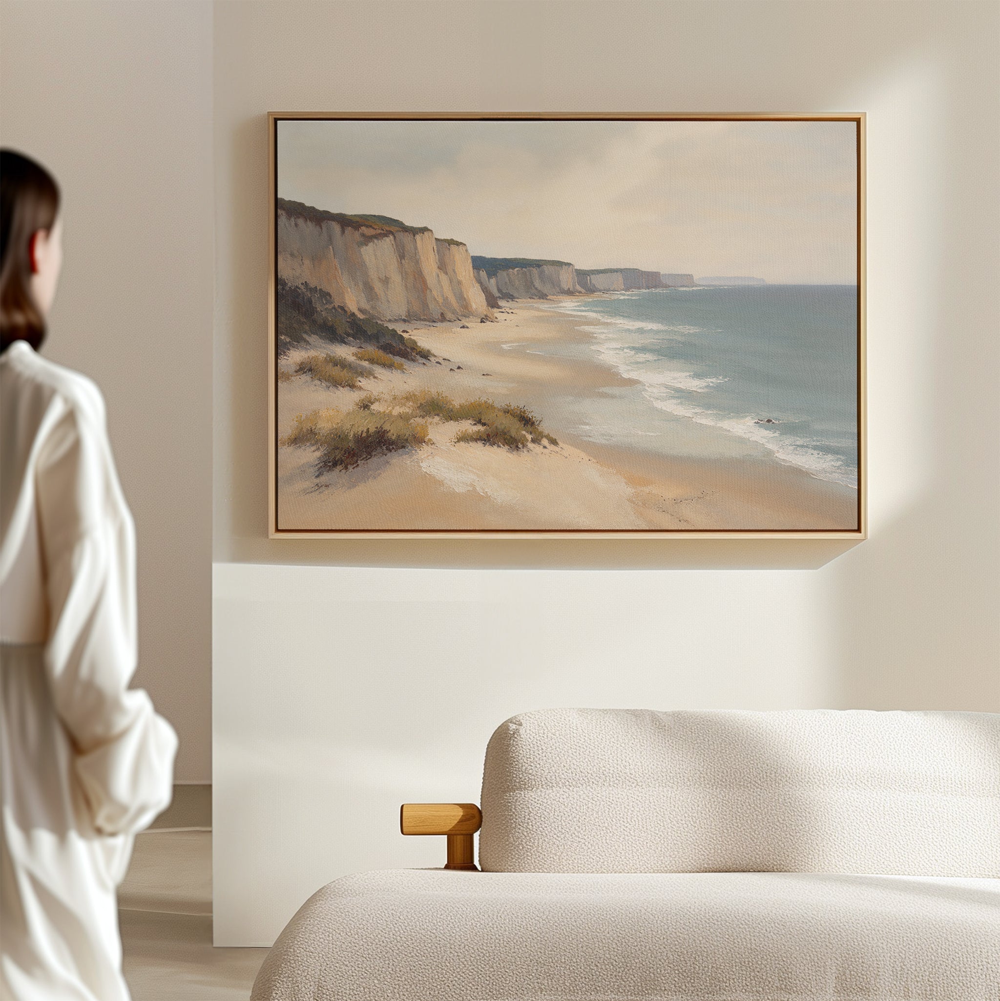 A woman admires the Chalk Shores Canvas Art - Horizontal, featuring sandy cliffs and gentle waves, hanging above a light-colored sofa in a softly lit room.