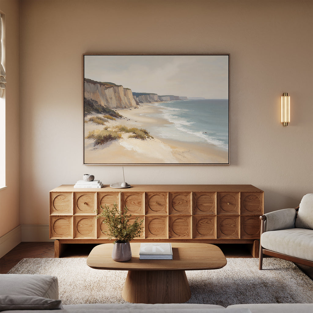 A cozy living room with neutral tones features a light sofa, wooden coffee table with a vase, patterned sideboard, and the Chalk Shores Canvas Art - Horizontal hanging above the sideboard for a seaside decor touch.