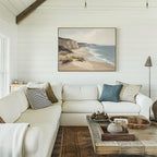 A cozy living room features a white sectional sofa, textured pillows, a rustic wooden coffee table, and the Chalk Shores Canvas Art - Horizontal above the couch. The space is finished with classic white shiplap walls.