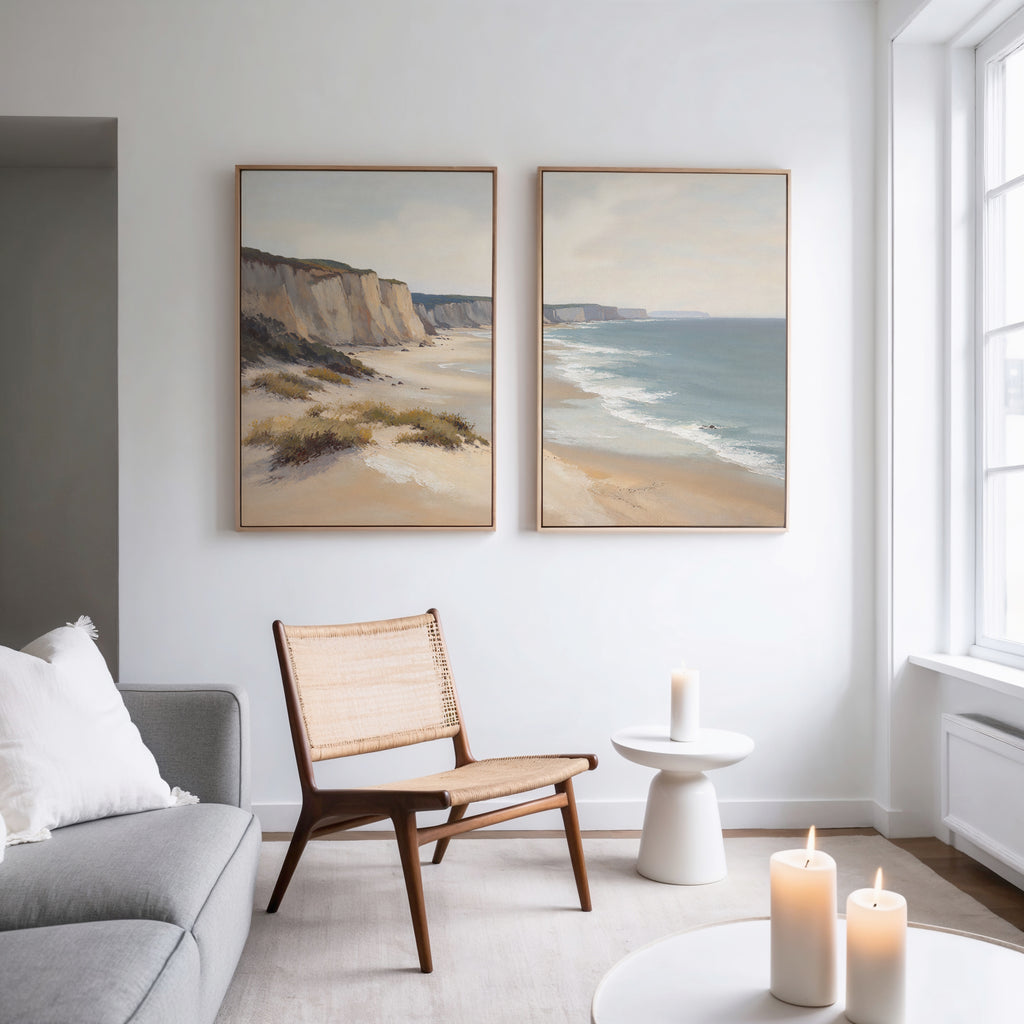 A bright, minimal living room features a gray sofa, woven chair, round table, and three lit candles. Above, the Chalk Shores - Set of 2 coastal landscape canvas prints add a serene touch to the white wall.