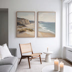 A bright, minimal living room features a gray sofa, woven chair, round table, and three lit candles. Above, the Chalk Shores - Set of 2 coastal landscape canvas prints add a serene touch to the white wall.