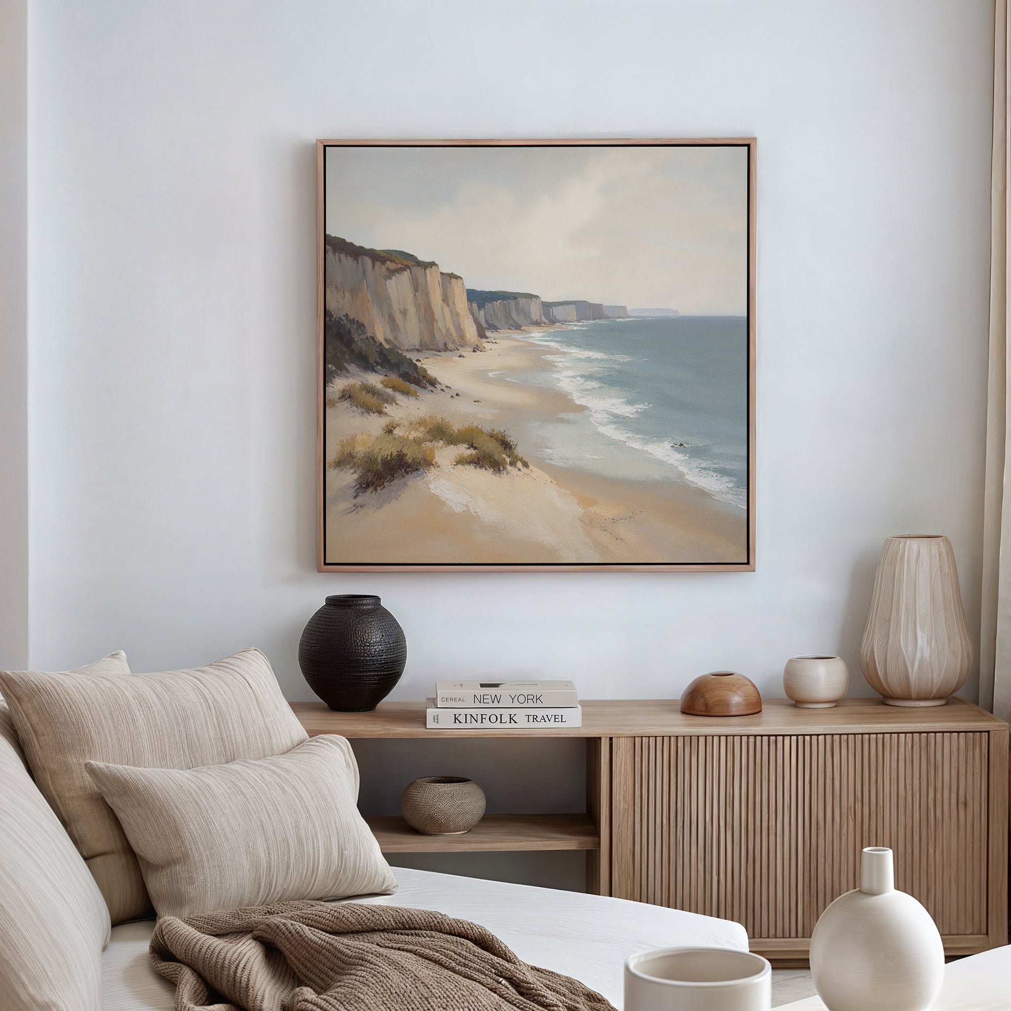 A cozy living room features a sofa with beige cushions, a wooden sideboard adorned with vases and books, and the Chalk Shores Canvas Art - Square depicting coastal cliffs and sea hanging on the white wall.