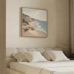 A minimal bedroom with a beige bed, white pillows, and the Chalk Shores Canvas Art - Square depicting sea cliffs and a beach hanging above the headboard on a light wall. A dried branch sits on the bedside table.