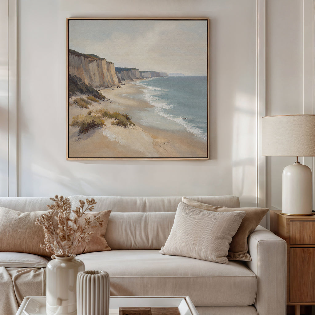 A bright, neutral-toned living room features a white sofa, beige pillows, and a wooden side table with a lamp. Above the sofa hangs the Chalk Shores Canvas Art - Square depicting a coastal scene. Dried flowers in a vase sit on the coffee table.