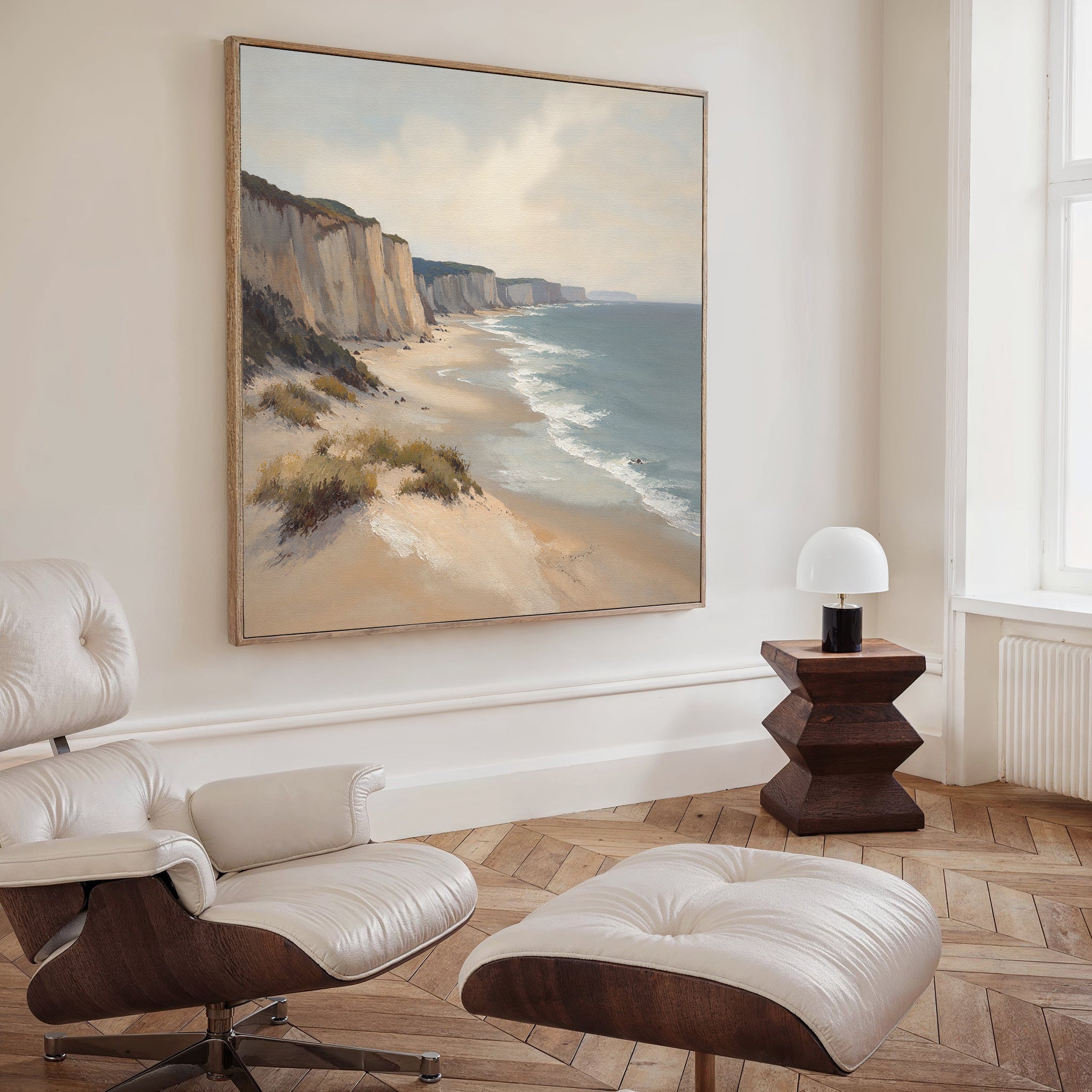A modern living room with a white lounge chair and ottoman, wooden side table with a white lamp, and the Chalk Shores Canvas Art - Square featuring cliffs and sandy beach, creating a tranquil seaside vibe.