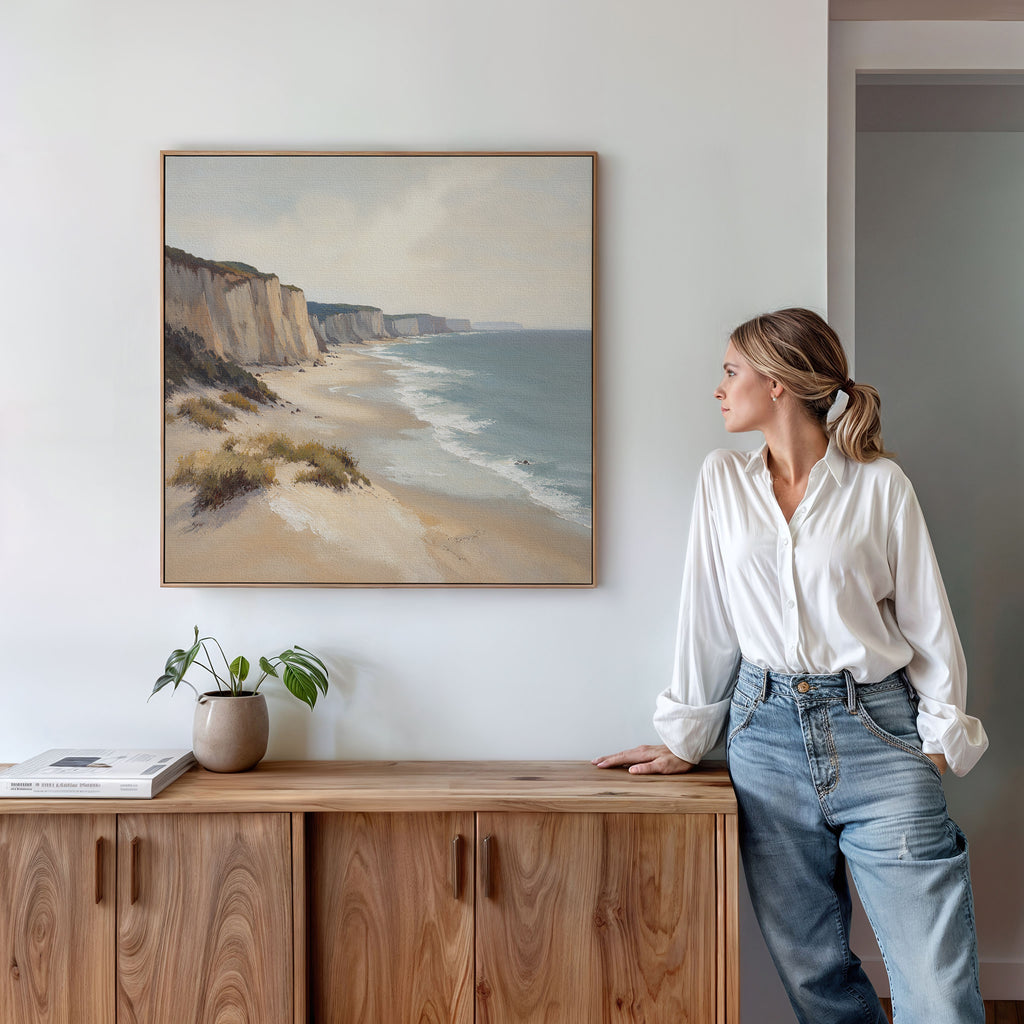 A woman in a white shirt and jeans stands by a wooden cabinet, admiring the Chalk Shores Canvas Art - Square depicting cliffs and sea, with a small plant and books nearby, creating a serene coastal setting.