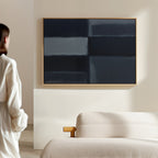 A person in white stands near a beige sofa, admiring the Charcoal Blocks Canvas Art - Horizontal with dark blue and black blocks on a light wall, showcasing modern living room decor.