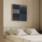 A minimalist bedroom features a beige upholstered bed with white bedding and four pillows. Above, the Charcoal Blocks Canvas Art - Square adds modern flair, while a bare branch decorates the bedside table.