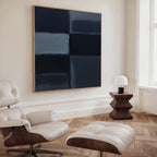 A modern, minimal living space featuring a white leather lounge chair with ottoman, a geometric dark wood side table with a white lamp, and the Charcoal Blocks Canvas Art - Square on a light wall—perfect for modern decor lovers.