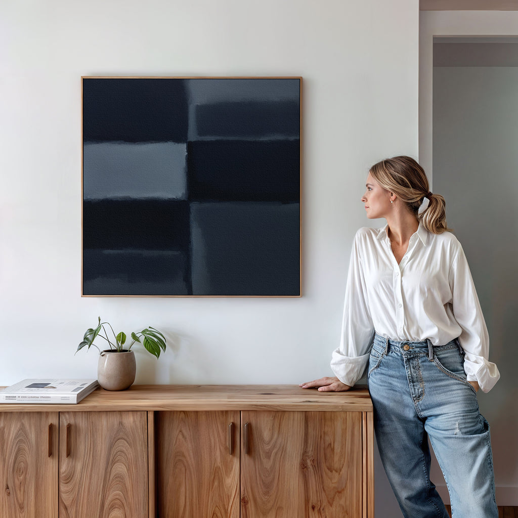 A woman in a white blouse and jeans leans on a wooden sideboard, admiring the Charcoal Blocks Canvas Art - Square. A potted plant and books sit on the sideboard, creating a minimalist, modern home decor atmosphere.