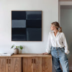 A woman in a white blouse and jeans leans on a wooden sideboard, admiring the Charcoal Blocks Canvas Art - Square. A potted plant and books sit on the sideboard, creating a minimalist, modern home decor atmosphere.