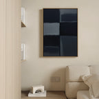 A minimal living room features a beige sofa, light wood shelves with books, and Charcoal Blocks Canvas Art - Vertical: a modern abstract painting with dark and blue squares in a gold frame on a cream wall; a geometric white decor piece sits on the lower shelf.