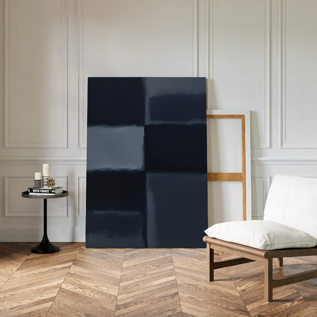 A minimalist room with wood herringbone flooring features a white cushioned chair, a round black side table with candles and books, and the Charcoal Blocks Canvas Art - Vertical leaning against a white paneled wall.
