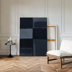A minimalist room with wood herringbone flooring features a white cushioned chair, a round black side table with candles and books, and the Charcoal Blocks Canvas Art - Vertical leaning against a white paneled wall.