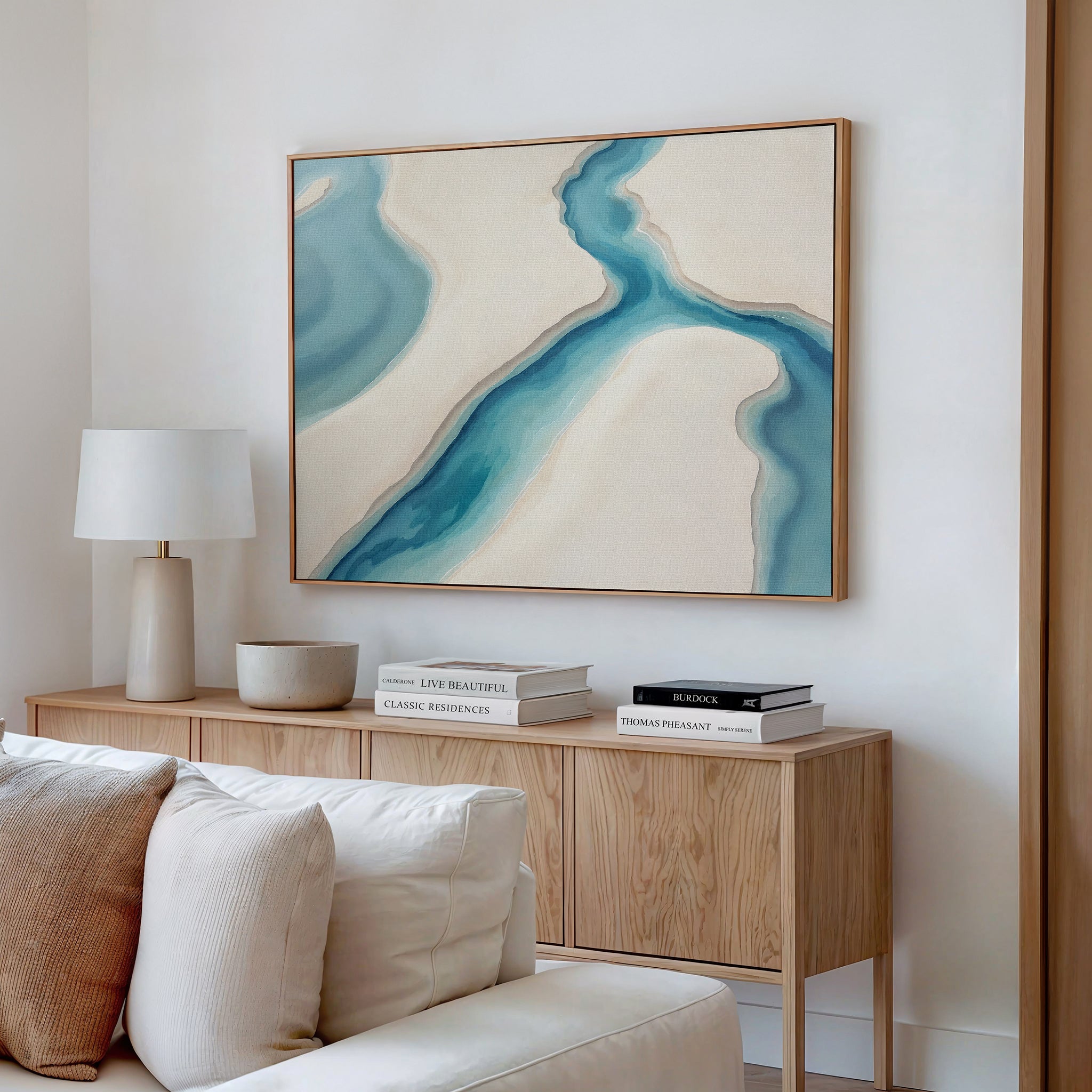A modern living room features a wooden sideboard with books, a lamp, and a bowl. Above hangs the Coastal Dreams Canvas Art - Horizontal in serene blue and beige tones. A white sofa with cushions sits in the foreground.