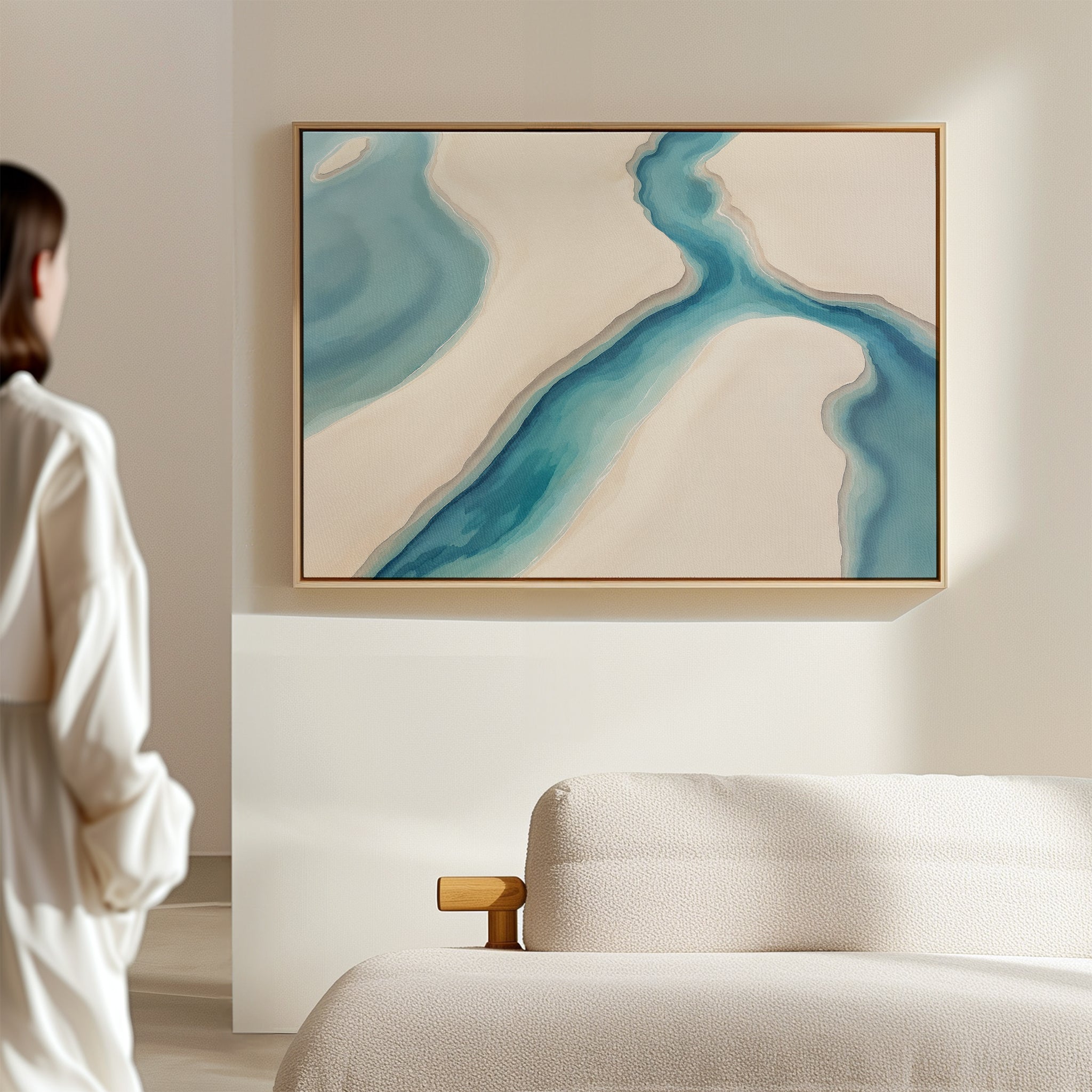 A woman in a white shirt admires the Coastal Dreams Canvas Art - Horizontal, featuring blue and beige shapes, displayed above a light-colored sofa in a minimalist modern room.