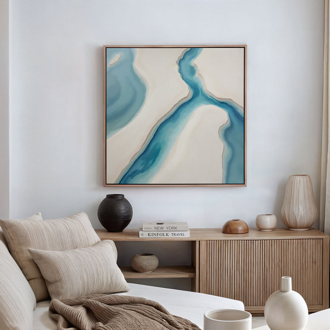 A cozy living space features a beige sofa with neutral cushions, a wooden sideboard with vases and books, and the Coastal Dreams Canvas Art - Square in blue and cream, adding tranquility to the white walls.