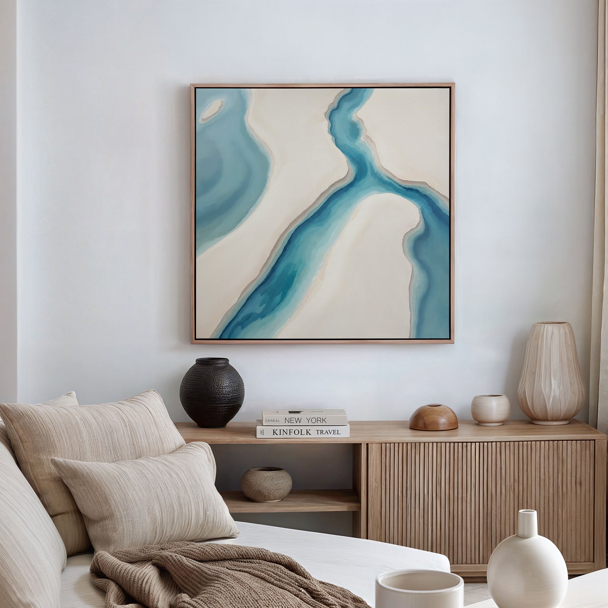 A cozy living space features a beige sofa with neutral cushions, a wooden sideboard with vases and books, and the Coastal Dreams Canvas Art - Square in blue and cream, adding tranquility to the white walls.