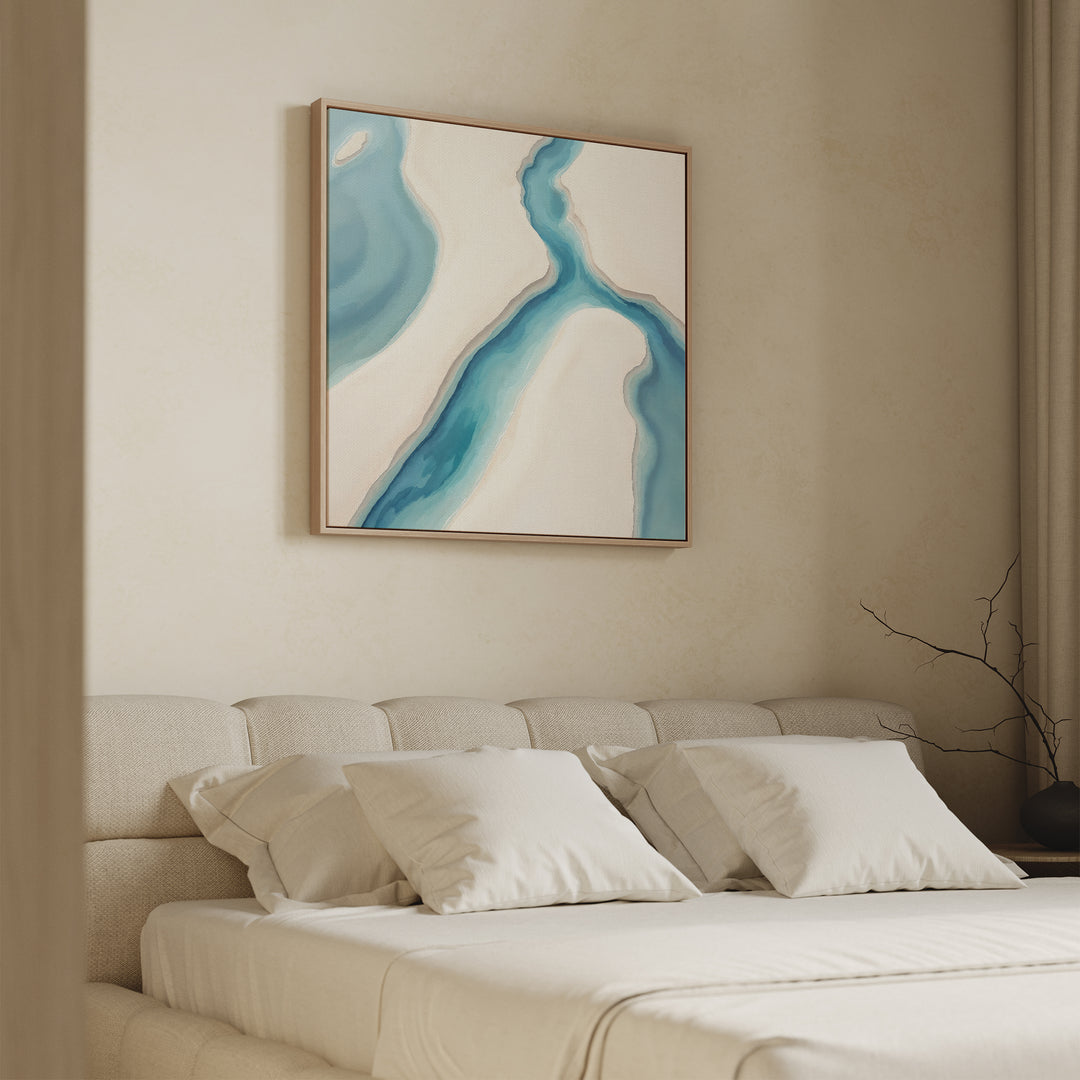 A modern bedroom features a beige upholstered bed with white bedding and pillows. The "Coastal Dreams Canvas Art - Square" in blue and white hangs above the bed, while a bare branch in a vase decorates the bedside table near soft curtains.