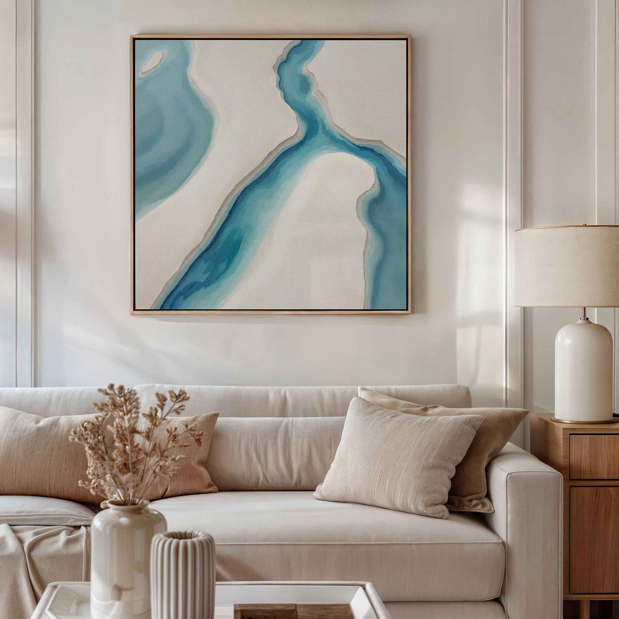 A modern living room with a beige sofa, light cushions, a wooden side table, cream lamp, dried flowers in a vase, and the Coastal Dreams Canvas Art - Square featuring blue and white shapes displayed above the couch.