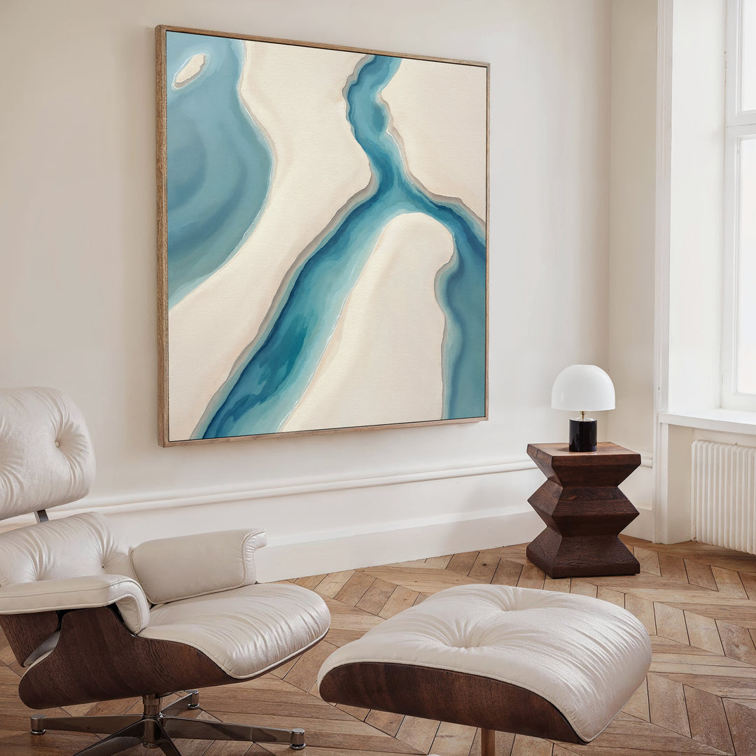 A modern living room featuring a cream leather lounge chair and ottoman, a wooden side table with a white lamp, and the Coastal Dreams Canvas Art - Square showcasing blue and beige shapes for a calming ambiance.