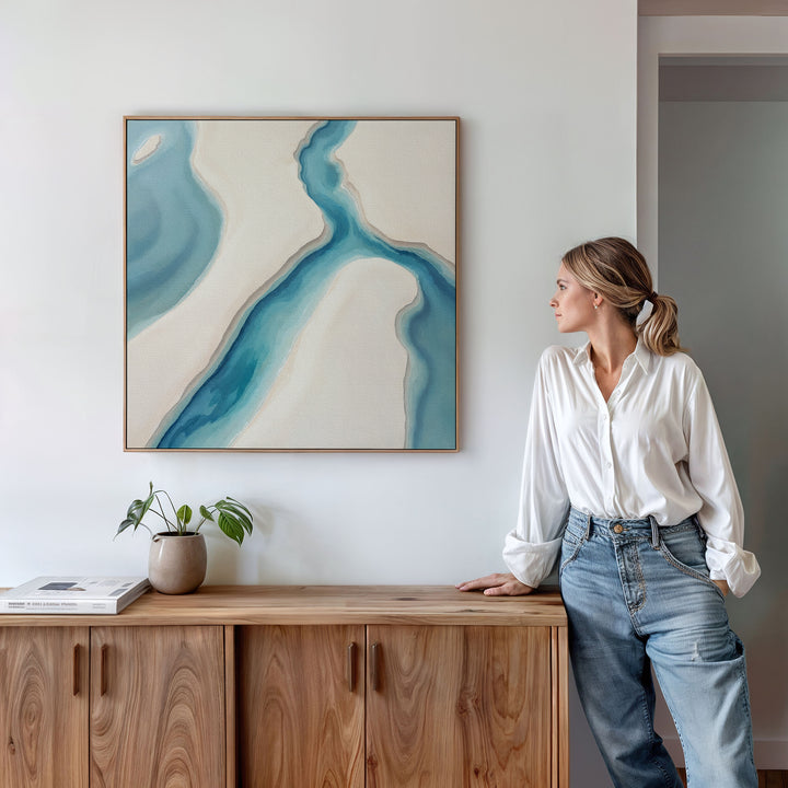 A woman in a white blouse and jeans leans against a wooden cabinet, admiring the Coastal Dreams Canvas Art - Square above. A potted plant and books on the cabinet enhance the calming, serene atmosphere.
