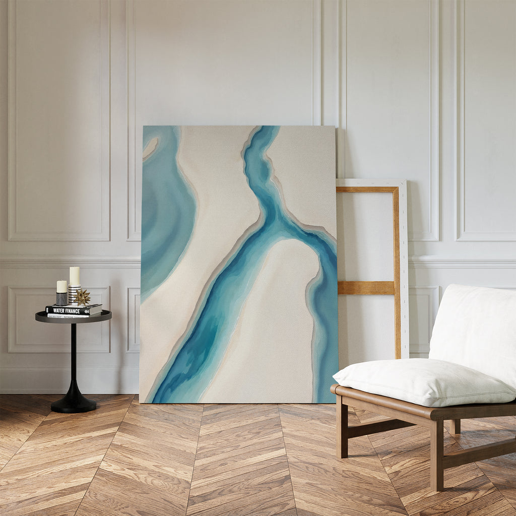 The Coastal Dreams Canvas Art - Vertical, featuring blue and beige river-like shapes, leans against a white-paneled wall beside a cushioned chair and black side table with candles on a herringbone wood floor.