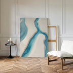 The Coastal Dreams Canvas Art - Vertical, featuring blue and beige river-like shapes, leans against a white-paneled wall beside a cushioned chair and black side table with candles on a herringbone wood floor.