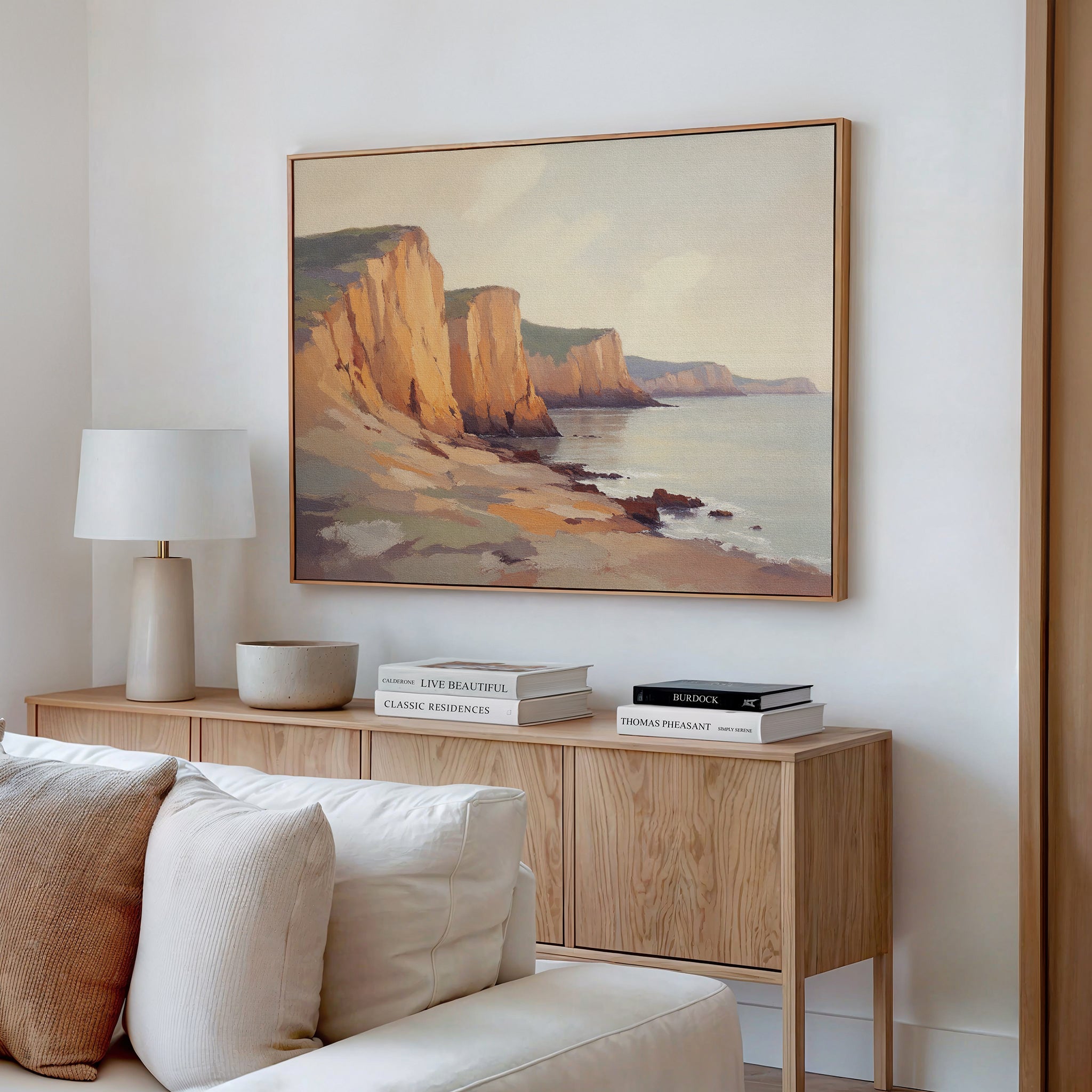 A minimalist living room features a light wood sideboard topped with a white table lamp and stacked books, while the Coastal Grandeur Canvas Art - Horizontal hangs above. A beige sofa in the foreground adds to the serene seaside charm.