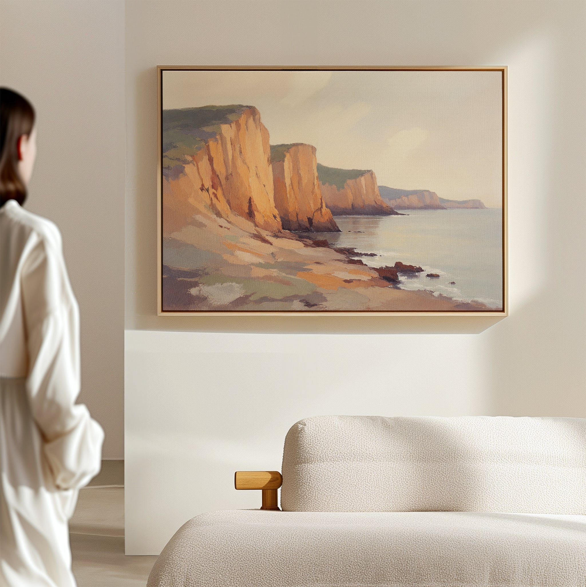 A person stands in a softly lit room, admiring the Coastal Grandeur Canvas Art - Horizontal featuring coastal cliffs and a calm sea, which is framed and hangs above a beige sofa on a light-colored wall.