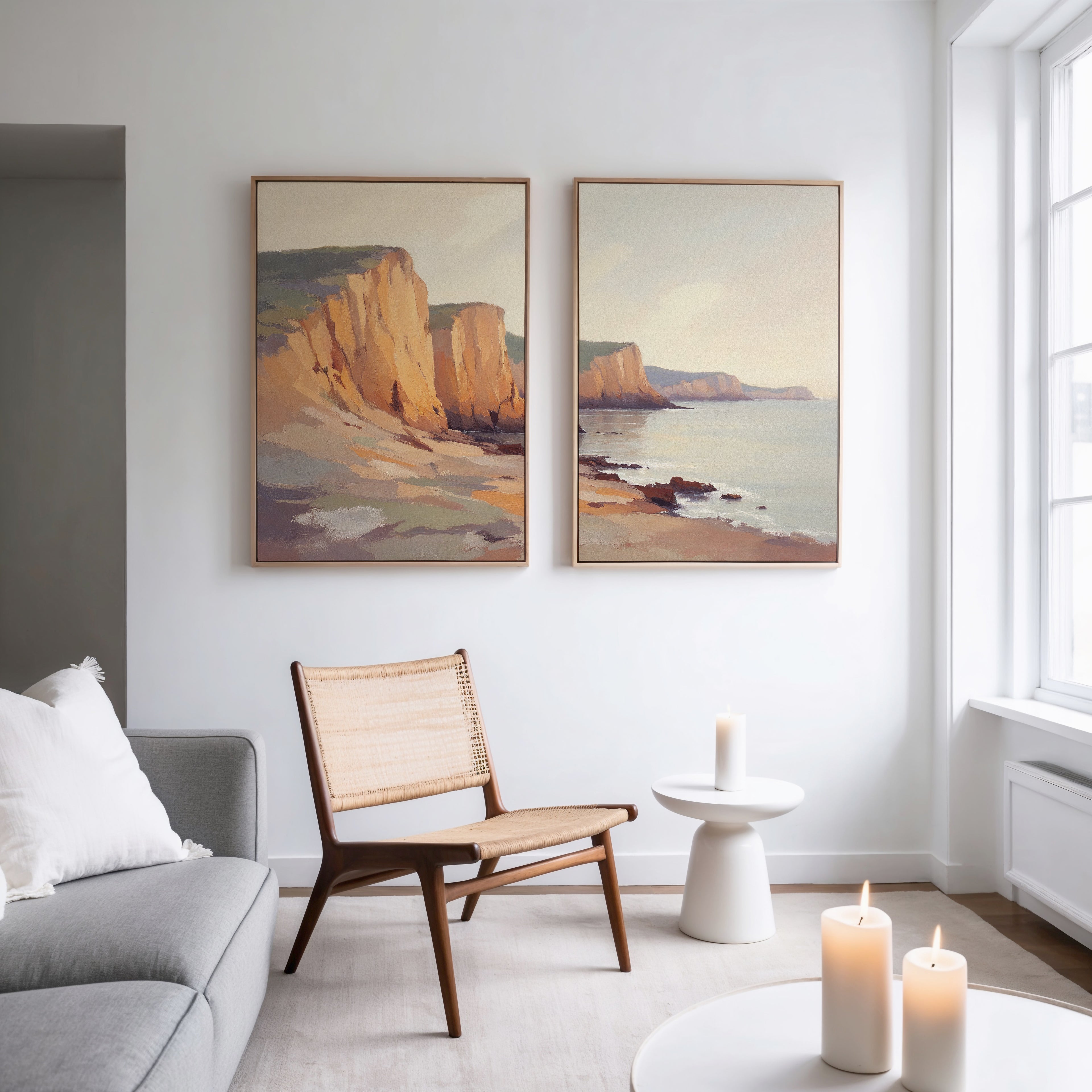 A modern, minimalist living room with a wooden chair, small white table, lit candles, gray sofa, and the Coastal Grandeur - Set of 2 canvas prints on a white wall by large windows creates a serene coastal atmosphere.