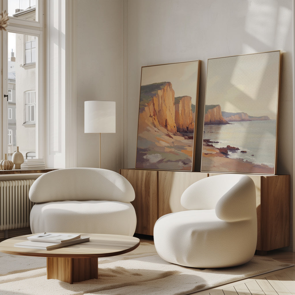 A modern living room features two white, curved armchairs, a wooden coffee table, and a sideboard displaying Coastal Grandeur - Set of 2 canvas prints. Sunlight pours in through large windows, creating a serene coastal vibe.