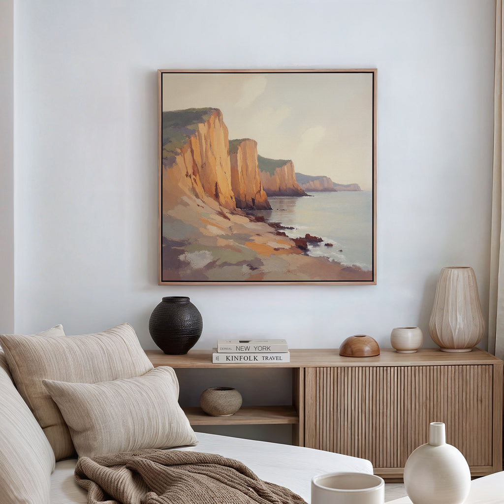 A cozy living room features a beige sofa, neutral cushions, a wooden sideboard with vases and books, and the Coastal Grandeur Canvas Art - Square—a framed painting of rocky seaside cliffs—hanging on the white wall above.