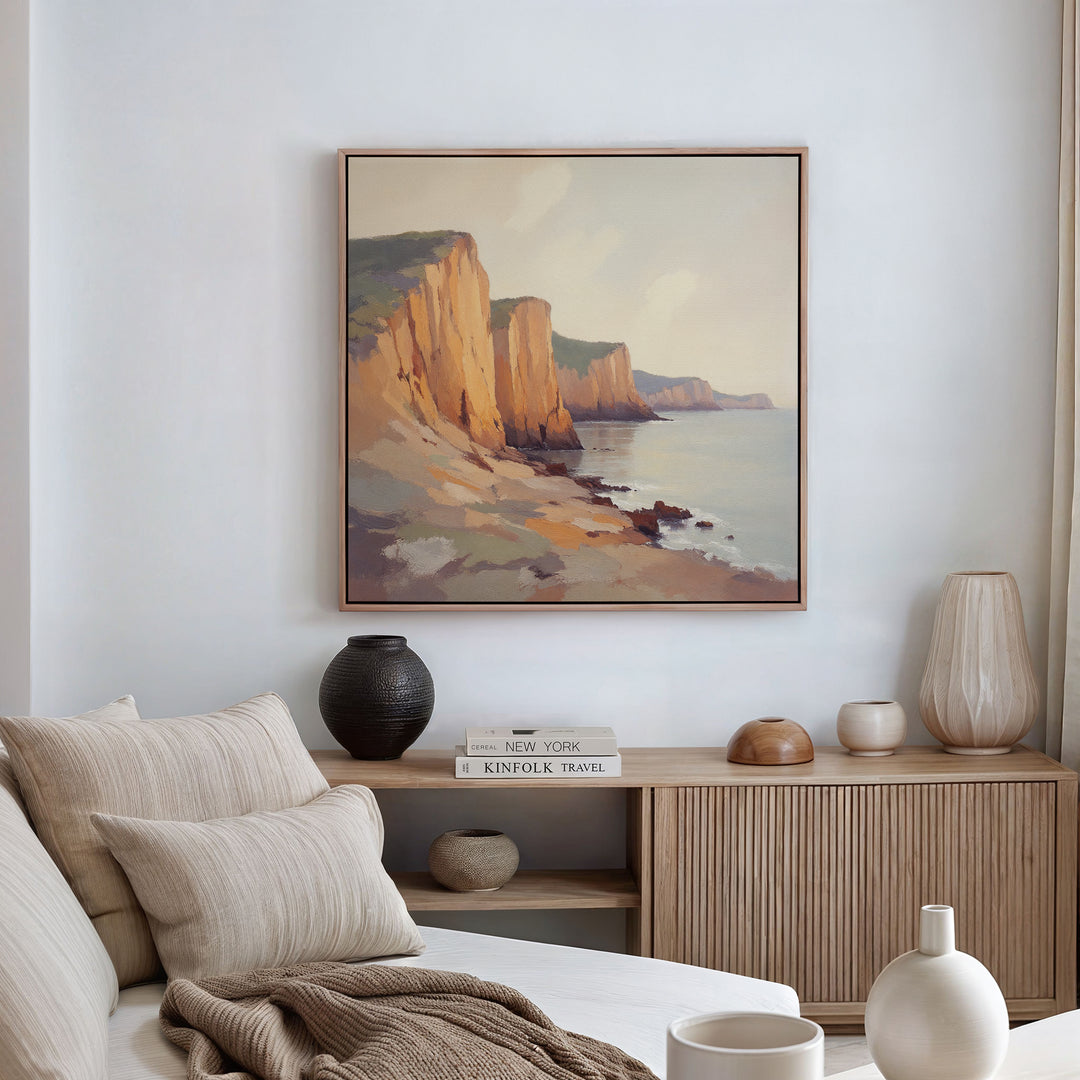 A cozy living room features a beige sofa, neutral cushions, a wooden sideboard with vases and books, and the Coastal Grandeur Canvas Art - Square—a framed painting of rocky seaside cliffs—hanging on the white wall above.