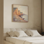 A cozy bedroom features a beige upholstered bed with white pillows and the Coastal Grandeur Canvas Art - Square, depicting seaside cliffs and sea, hanging above the headboard. Soft natural light complements the neutral tones.