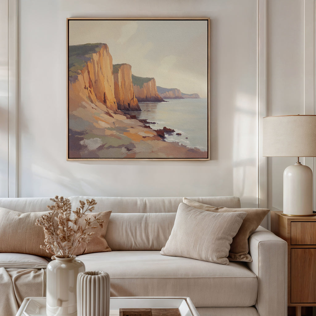 A living room with a beige sofa, neutral cushions, a side table with a lamp, and a vase of dried flowers. Above the sofa hangs the Coastal Grandeur Canvas Art - Square featuring seaside cliffs, giving the room a warm, minimalist vibe.