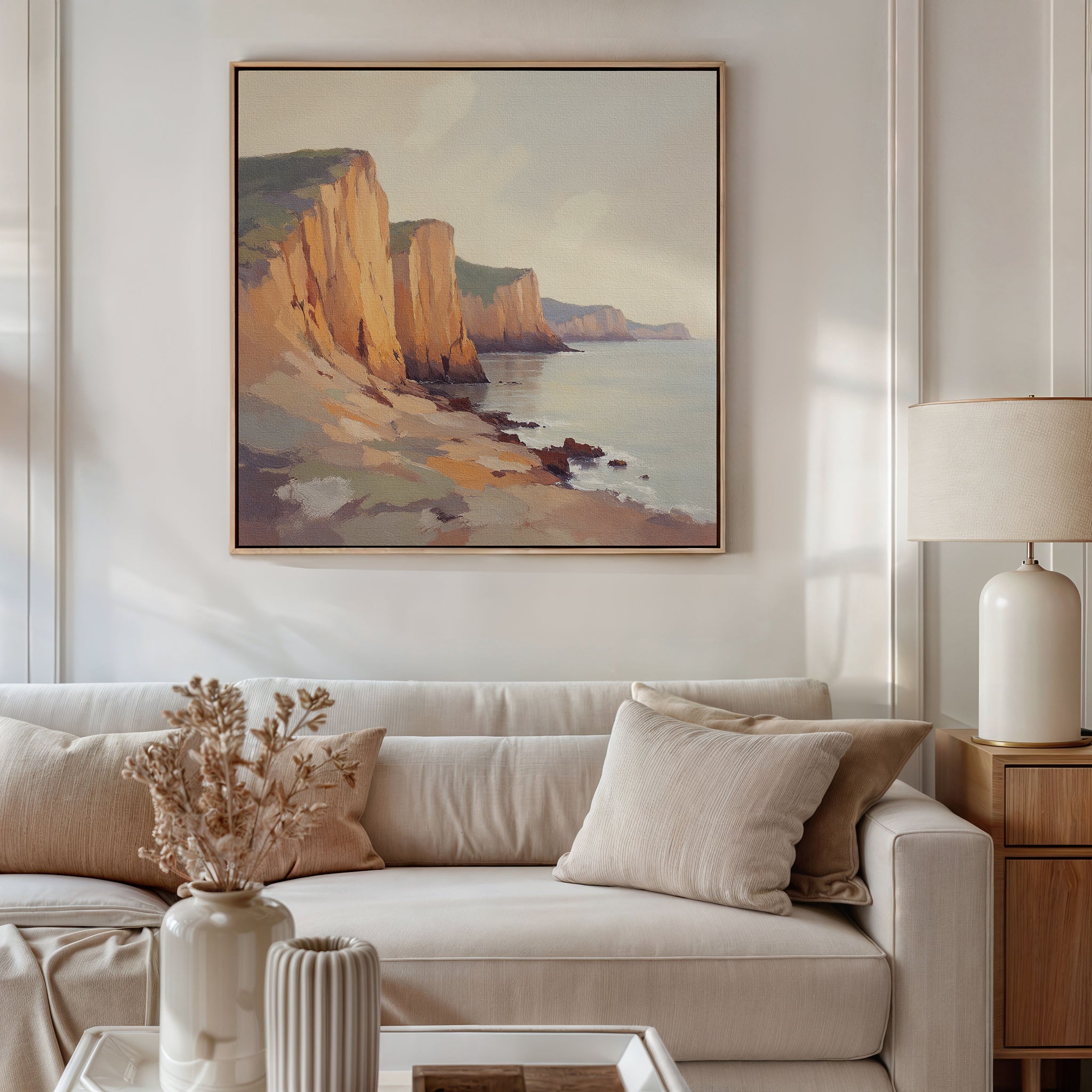 A living room with a beige sofa, neutral cushions, a side table with a lamp, and a vase of dried flowers. Above the sofa hangs the Coastal Grandeur Canvas Art - Square featuring seaside cliffs, giving the room a warm, minimalist vibe.