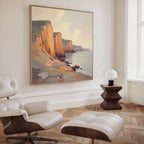 A modern living room with a white leather lounge chair and ottoman, wooden side table with a small lamp, and the Coastal Grandeur Canvas Art - Square featuring rocky seaside cliffs on a beige wall.