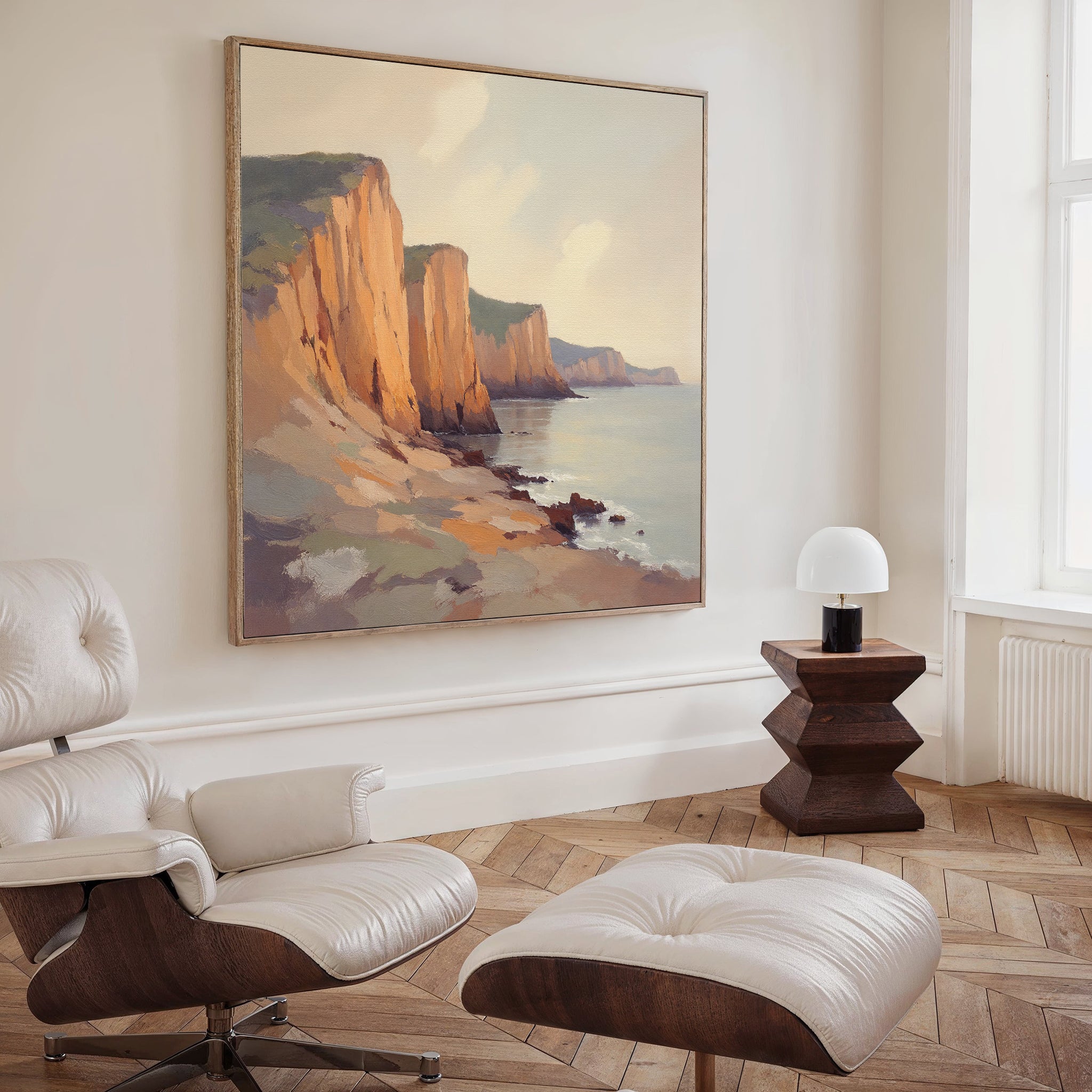 A modern living room with a white leather lounge chair and ottoman, wooden side table with a small lamp, and the Coastal Grandeur Canvas Art - Square featuring rocky seaside cliffs on a beige wall.