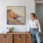 A woman in a white blouse and jeans stands by a wooden cabinet, gazing at the "Coastal Grandeur Canvas Art - Square"—a framed seaside cliff print—hanging on the white wall above her. A potted plant and a book rest on the cabinet.