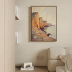The Coastal Grandeur Canvas Art - Vertical, featuring seaside cliffs at sunset, hangs on a beige wall above a light sofa. A built-in shelf with books and a small white decor piece is partially visible to the left.
