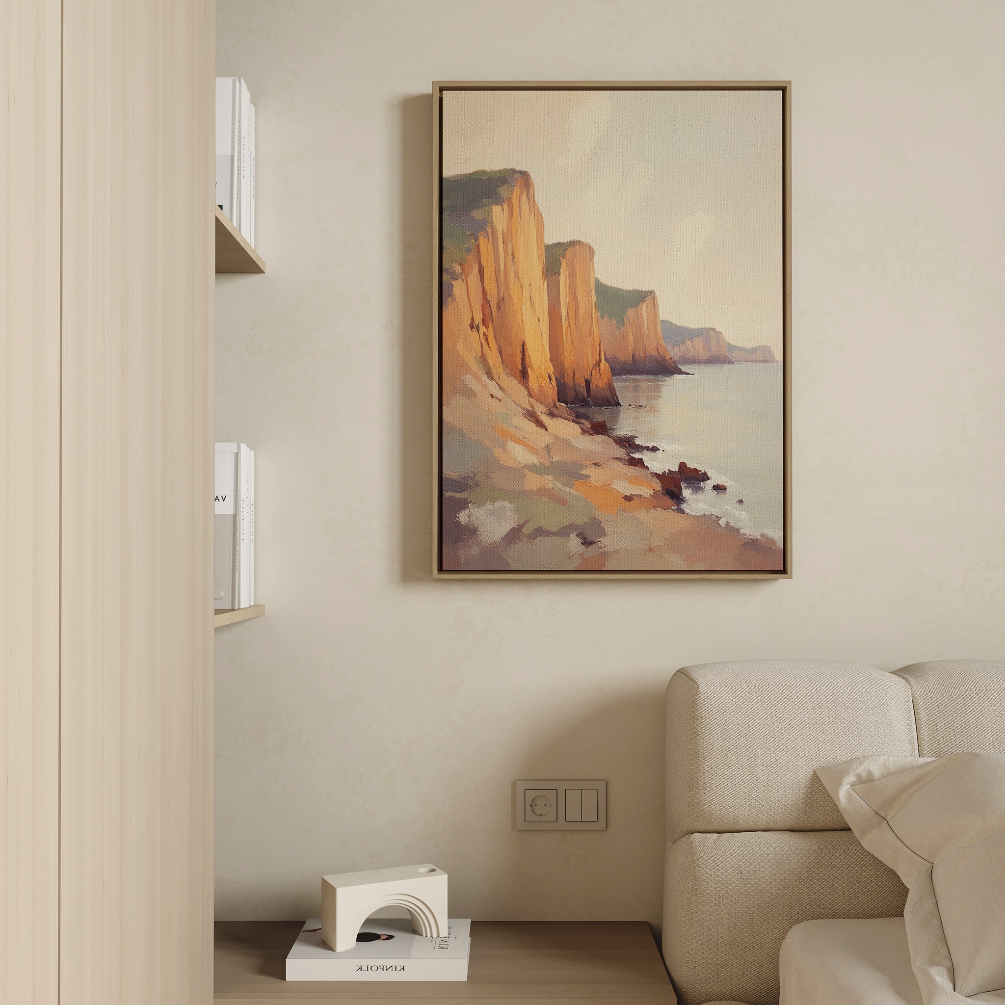 The Coastal Grandeur Canvas Art - Vertical, featuring seaside cliffs at sunset, hangs on a beige wall above a light sofa. A built-in shelf with books and a small white decor piece is partially visible to the left.