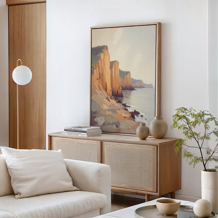 A modern living room in neutral tones featuring a wooden sideboard, ceramic vases, books, a potted plant, and the Coastal Grandeur Canvas Art - Vertical depicting seaside cliffs hanging above the sideboard.