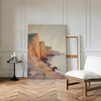 The Coastal Grandeur Canvas Art - Vertical rests against a white paneled wall in a minimalist room, beside an empty frame. Nearby, a cushioned chair and small round table with candles and books sit on the wooden herringbone floor.