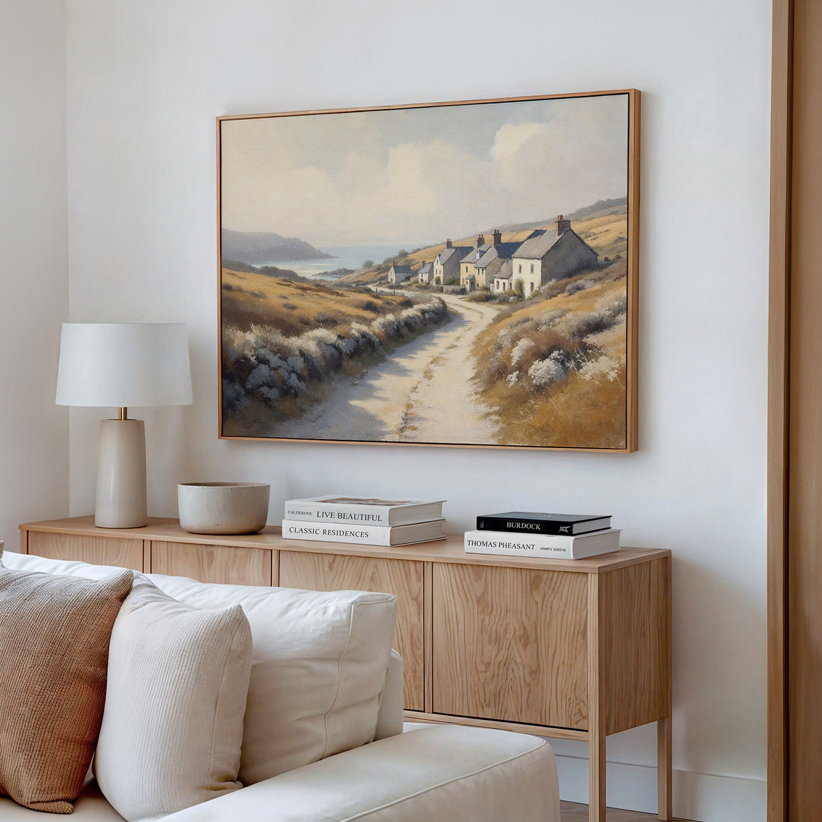 A cozy living room features a light wood console table with books, a lamp, and a bowl. Above it hangs the Coastal Haven Canvas Art - Horizontal, showcasing cottages by the sea. A white sofa sits in the foreground.